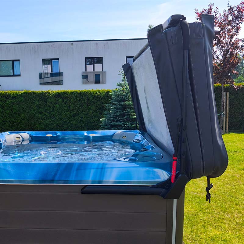 Basseini kaanetõstuk CoverMate III | Santeco Kotikylpylä Blue outdoor hot tub with the insulating cover folded up on the side in a sunny backyard with a hedge and building in the background