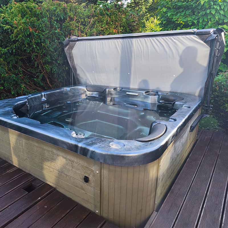 Basseini kattetõstuk ALifter III5 | Santeco Kotikylpylä Outdoor hot tub on a wooden deck with its cover raised filled with water and built in jets surrounded by greenery